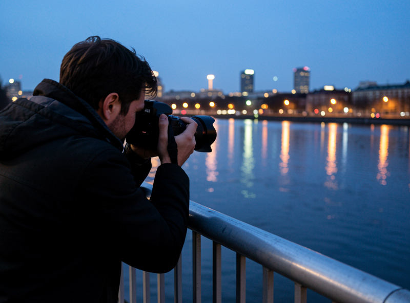 A photographer bracing a camera against a railing to shoot a blue-hour cityscape with glowing lights reflected in water, hand-held stabilization technique