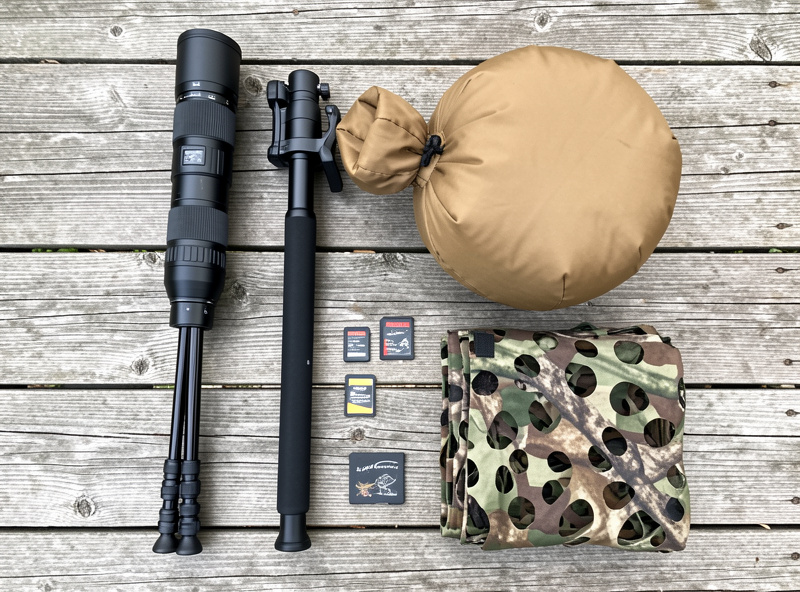 Flat-lay of wildlife photography kit including a long telephoto zoom, monopod, bean bag, memory cards and camouflage netting