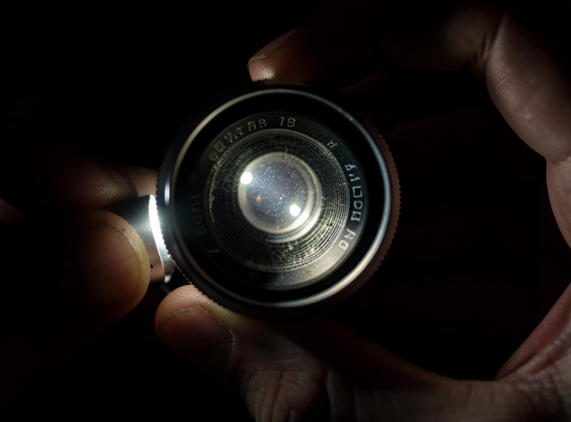 Inspecting a used vintage lens with a flashlight to check for internal fungus and haze against a dark background