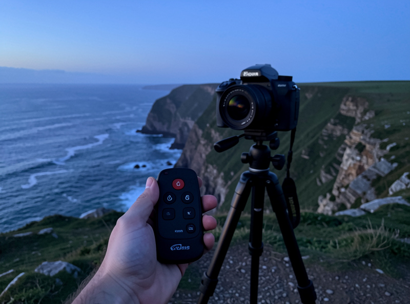 A wireless shutter remote held in a photographer's hand next to a tripod-mounted camera on a coastal cliff at blue hour
