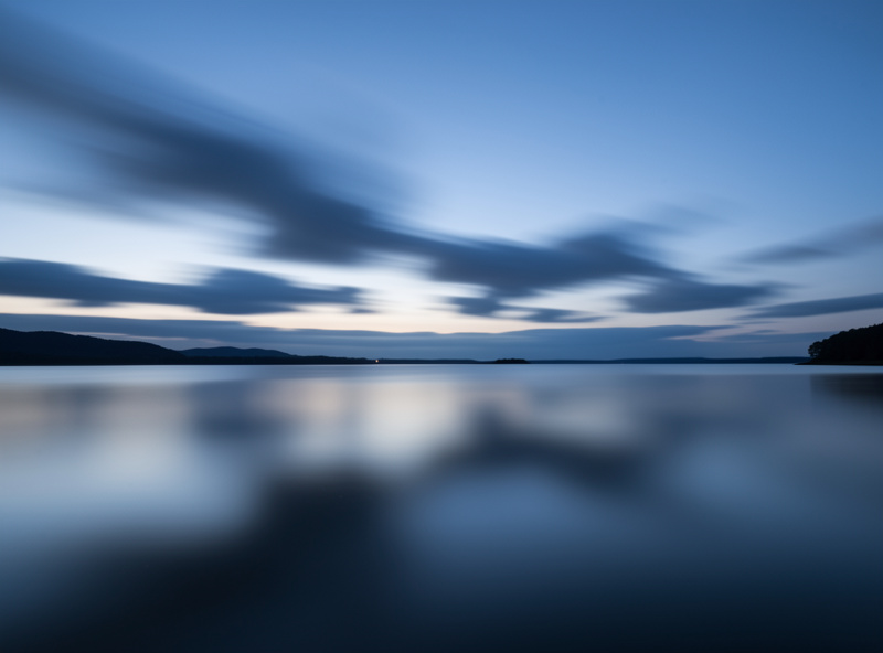 Long exposure of soft blurred clouds streaking sideways above a calm reflective lake at blue hour