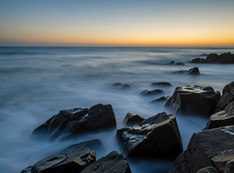Coastal seascape long exposure at dusk with waves smoothed into low mist around dark foreground rocks