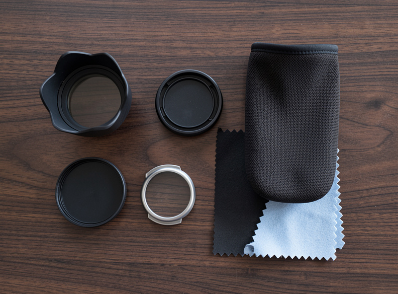 Flat lay of a petal lens hood, front and rear caps, a padded lens pouch, and a microfiber cloth on a dark wood surface