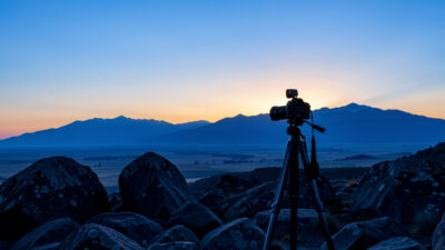 Tripod-mounted camera on a rocky foreground with sharp boulders and a distant ridge at blue-hour dawn