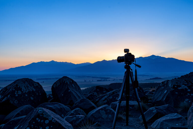 Tripod-mounted camera on a rocky foreground with sharp boulders and a distant ridge at blue-hour dawn