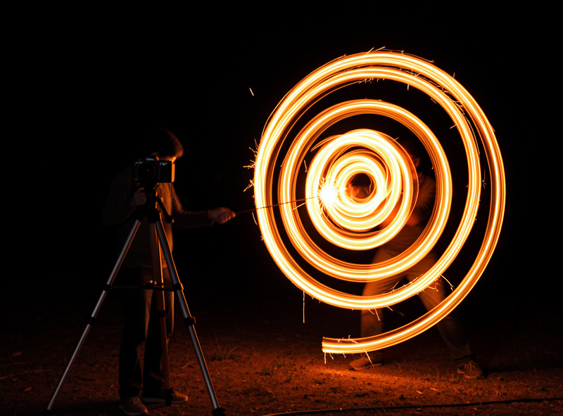 A camera on a tripod in the dark pointed at a person drawing a spiral of orange light with a sparkler