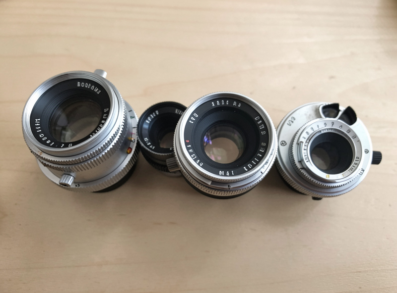 A group of M42 screw-mount vintage prime lenses arranged on a table with a mirrorless camera